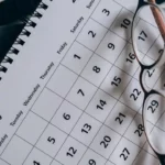 Un gros plan sur un calendrier relié en spirale affichant le mois de janvier, posé sur une surface sombre, avec des lunettes reposant en diagonale dessus.